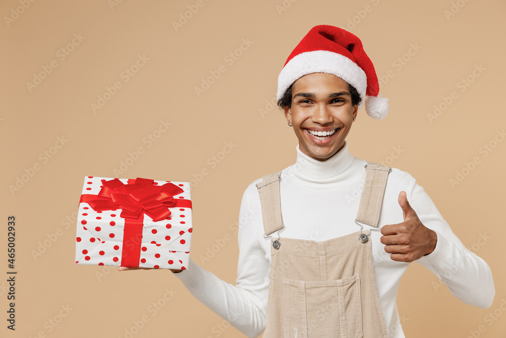 Young african man 20s wear Santa Claus red Christmas hat hold present box with gift ribbon bow sho thumb up isolated on plain pastel beige background studio. Happy New Year 2022 celebration concept. Young african man 20s wear Santa Claus red Christmas hat hold present box with gift ribbon bow sho thumb up isolated on plain pastel beige background studio. Happy New Year 2022 celebration concept.