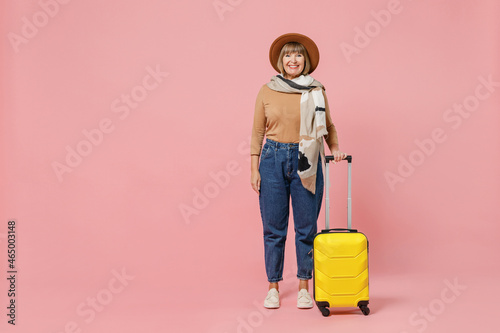 Full size body length traveler tourist vivid mature elderly senior woman 55 years old wears casual clothes hat scarf hold suitcase bag isolated on plain pastel light pink background studio portrait.