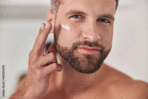 Attractive mature man applies cream onto skin under eye in bathroom