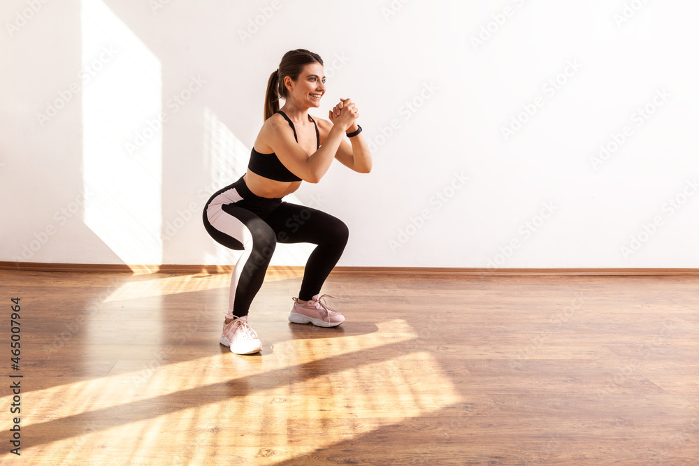Fototapeta premium Profile of athletic woman doing squat, lower body sport exercise, warming up and training muscles, wearing black sports top and tights. Full length studio shot illuminated by sunlight from window.