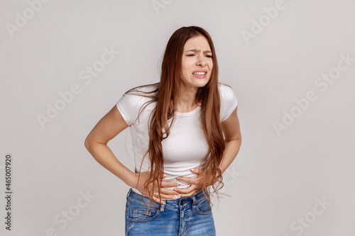 Portrait of unhealthy distressed woman clutching her belly, grimacing from acute abdominal pain, gastritis or constipation, wearing white T-shirt. Indoor studio shot isolated on gray background.