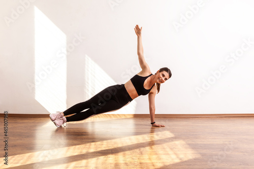 Fitness girl practicing yoga, doing Vasisthasana exercise with one hand raised, training muscles, wearing black sports top and tights. Full length studio shot illuminated by sunlight from window.