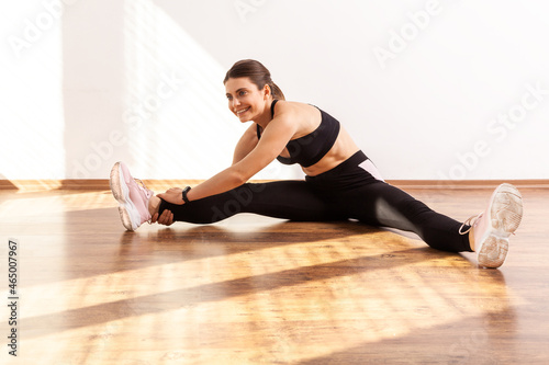 Flexible woman sits in legs split, bending to touch feet, stretching muscle, flexibility exercises, wearing black sports top and tights. Full length studio shot illuminated by sunlight from window.