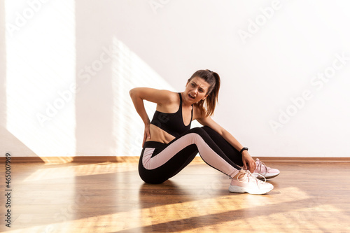 Athletic female frowning from pain in lower back, muscle strains and ligament sprains problem, wearing black sports top and tights. Full length studio shot illuminated by sunlight from window.