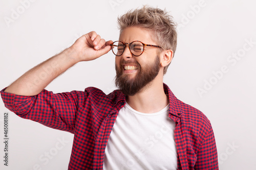 Portrait of funny extremely happy bearded man touching eyeglasses standing with closed eyes and laughing. Indoor studio shot isolated on gray background