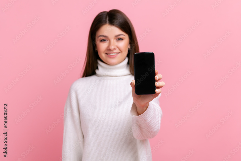 Smiling brunette female showing smartphone with blank screen for advertisement or promotional text, wearing white casual style sweater. Indoor studio shot isolated on pink background. Smiling brunette female showing smartphone with blank screen for advertisement or promotional text, wearing white casual style sweater. Indoor studio shot isolated on pink background.