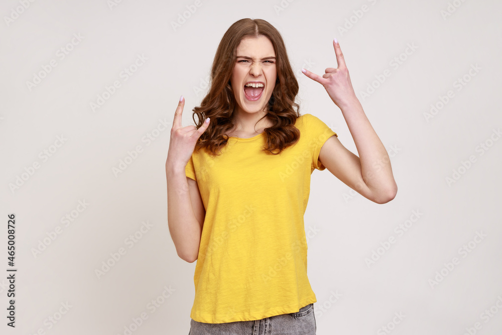 Extremely excited satisfied teenager girl with brown hair in yellow T-shirt showing rock and roll gesture with fingers, having fun, resting. Indoor studio shot isolated on gray background. Extremely excited satisfied teenager girl with brown hair in yellow T-shirt showing rock and roll gesture with fingers, having fun, resting. Indoor studio shot isolated on gray background.