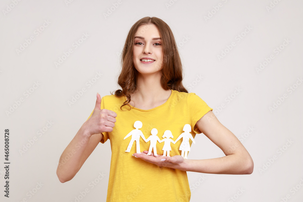 Portrait of happy satisfied female of young age holding paper family chain and showing thumb up, looking at camera with toothy smile. Indoor studio shot isolated on gray background. Portrait of happy satisfied female of young age holding paper family chain and showing thumb up, looking at camera with toothy smile. Indoor studio shot isolated on gray background.