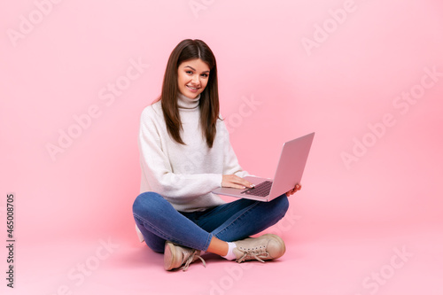 Positive satisfied female sitting with crossed legs on floor, holding laptop, looking at camera, wearing white casual style sweater. Indoor studio shot isolated on pink background.