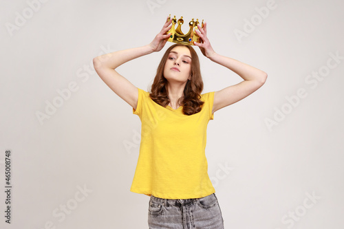 Obraz na plátně Portrait of selfish brown haired teenage girl in yellow T-shirt holding gold crown over head, having arrogance expression, privileged status
