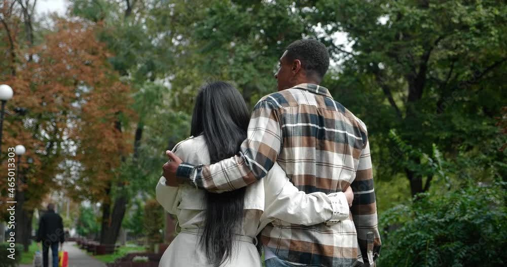 View from the back of a young man in checkered shirt and his girlfriend in beige trench coat hugging each other while walking on the street of a city park, having fun together at romantic date. View from the back of a young man in checkered shirt and his girlfriend in beige trench coat hugging each other while walking on the street of a city park, having fun together at romantic date.