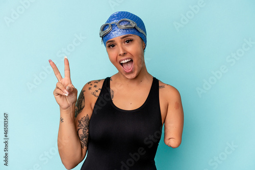 Young swimmer caucasian woman with one arm isolated on blue background joyful and carefree showing a peace symbol with fingers. photo