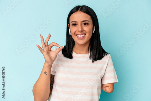 Young caucasian woman with one arm listening to music with wireless headphones isolated on blue background cheerful and confident showing ok gesture.