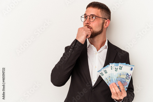 Young business caucasian man holding banknotes isolated on white background looking sideways with doubtful and skeptical expression.