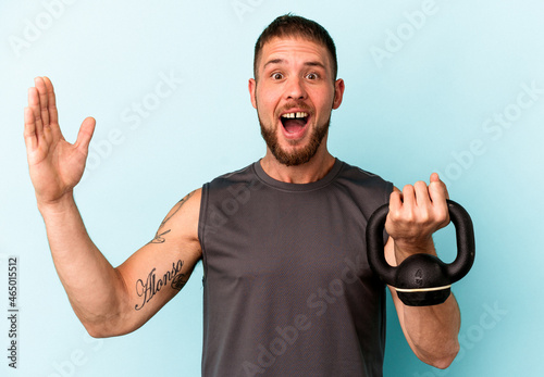 Young caucasian man holding ketlebell isolated on blue background receiving a pleasant surprise, excited and raising hands. photo