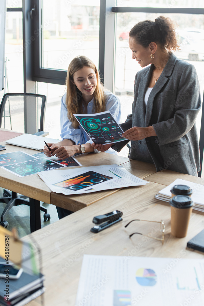 Obraz premium Smiling african american businesswoman holding paper near cheerful colleague near coffee to go and documents in office