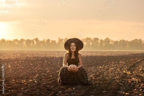 beautiful woman in a witch costume holds a pumpkin. witch in a black hat with a carved pumpkin stands in a field against background of the sky, sunset