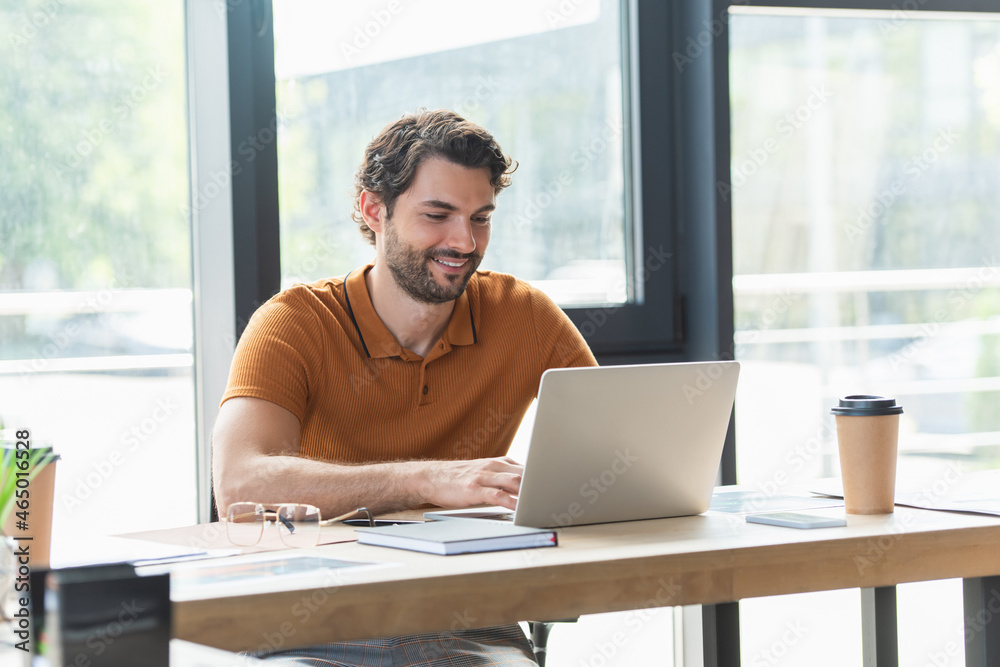 Smiling businessman using laptop near coffee and eyeglasses on table in office Smiling businessman using laptop near coffee and eyeglasses on table in office