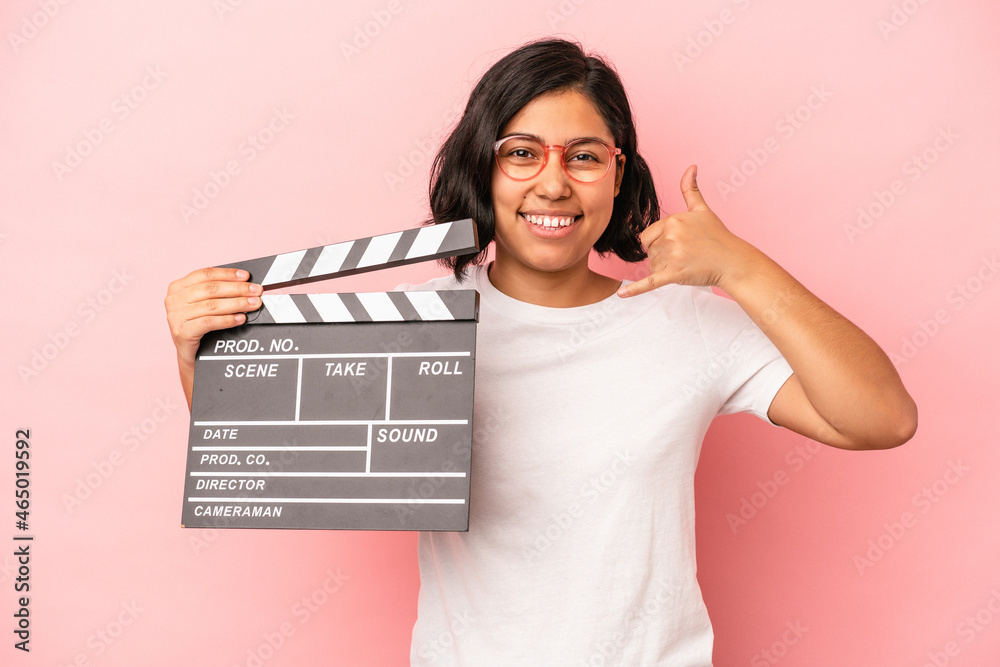 Young latin woman holding clapperboard isolated on pink background showing a mobile phone call gesture with fingers.