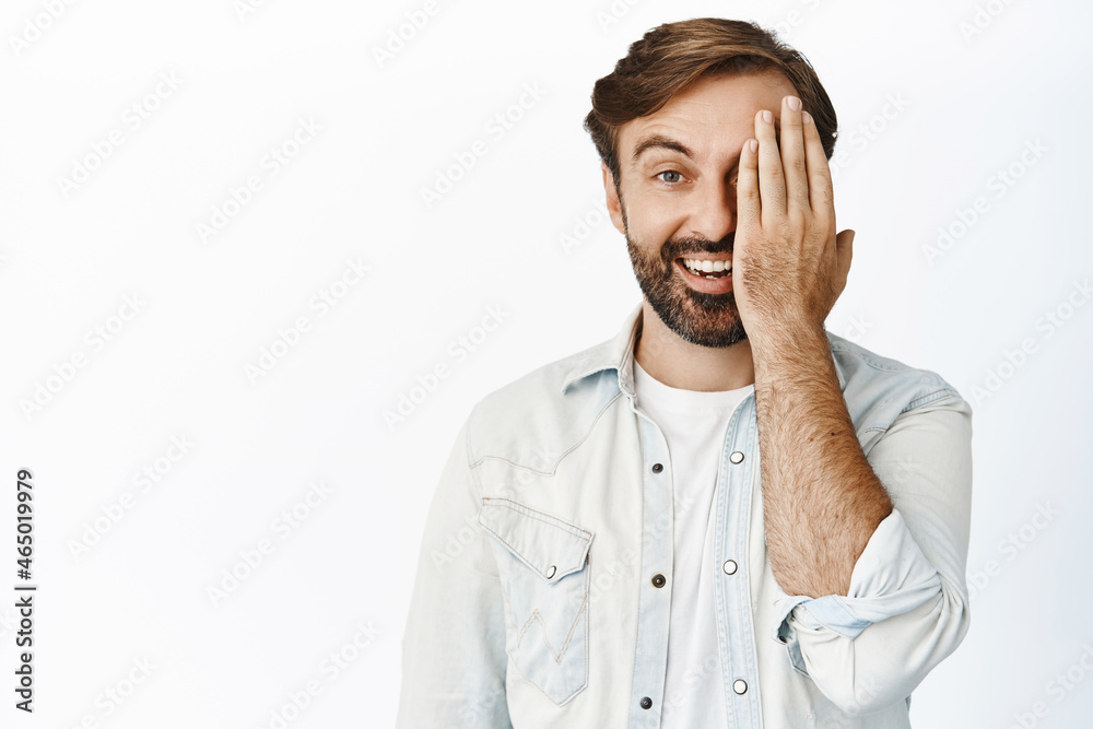 Portrait of happy guy cover half of face and smiling, looking with one side eye at camera, white background Portrait of happy guy cover half of face and smiling, looking with one side eye at camera, white background