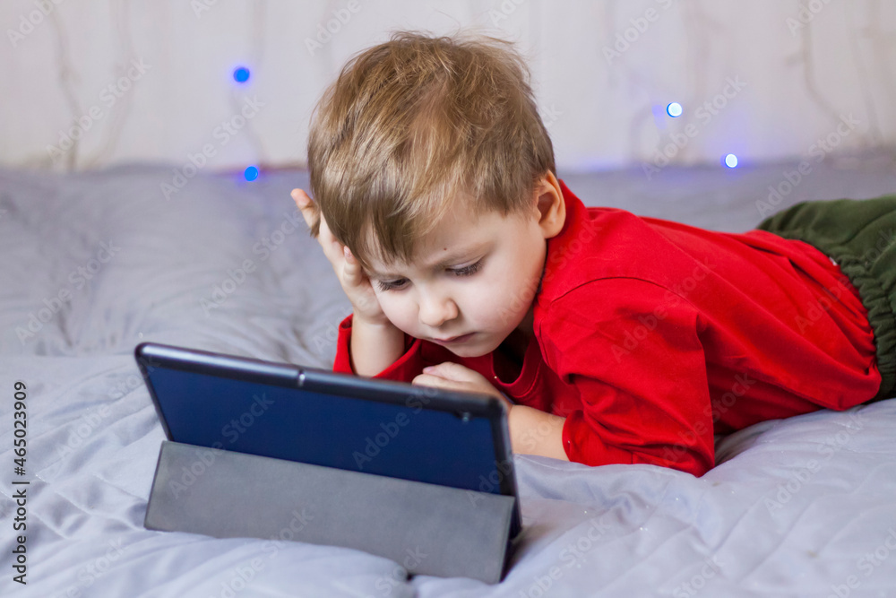 A cute boy is sitting on the bed and looking at a tablet. Funny smiling child. A cute boy is sitting on the bed and looking at a tablet. Funny smiling child.