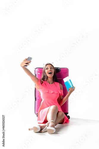 Emotional girl tourist at airport with pink suitcase  phone and boarding pass. Girl traveler isolated on white background. Travel concept. Girl in a pink mini dress at the airport.