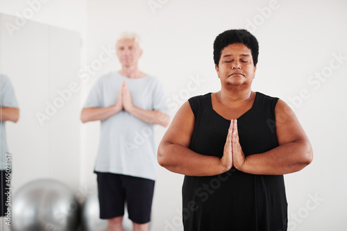 Senior people standing with eyes closed in yoga pose and meditating in fitness studio