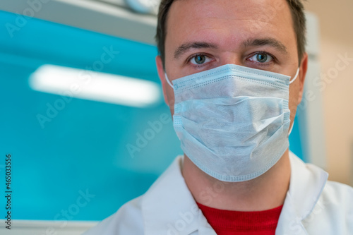 Portrait of scientist in medical mask in room with laboratory box in UV light. Tired successful male worker of scientific laboratory sitting by laboratory shelf inside office in front of camera