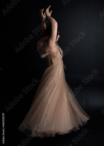 Full length portrait of red haired girl wearing a creamy fantasy gown like a fairy goddess costume. standing pose with elegant gestural movement backwards the the camera, isolated on dark studio 