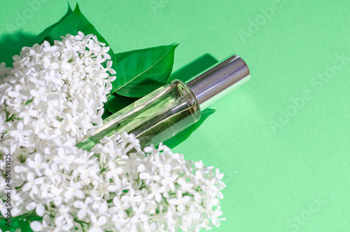 White lilac flowers and a glass bottle with perfume on a green background. Bright shadows and light. Place for your text. Flat lay.