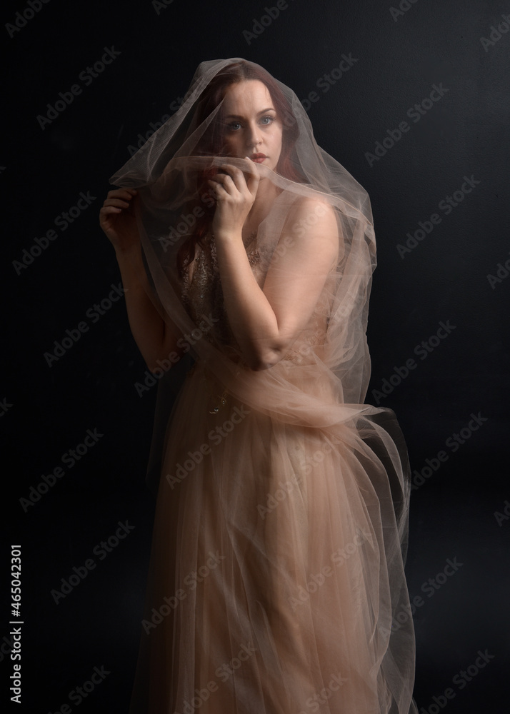 Full length  portrait of red haired  girl wearing a creamy fantasy gown like a fairy goddess costume.  standing  pose with elegant gestural movement , isolated on dark studio background.