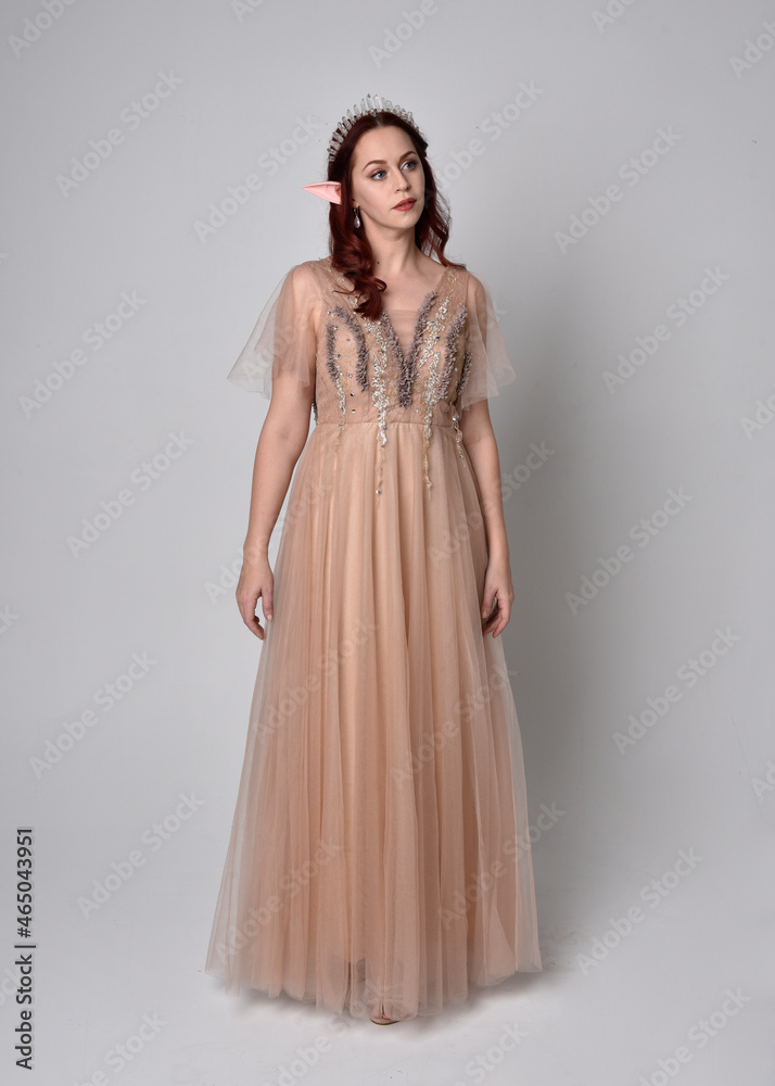 Full length  portrait of red haired  girl wearing a creamy fantasy gown and crystal crown, like a fairy goddess costume.  standing  pose with elegant gestural arm movement , isolated on light studio b