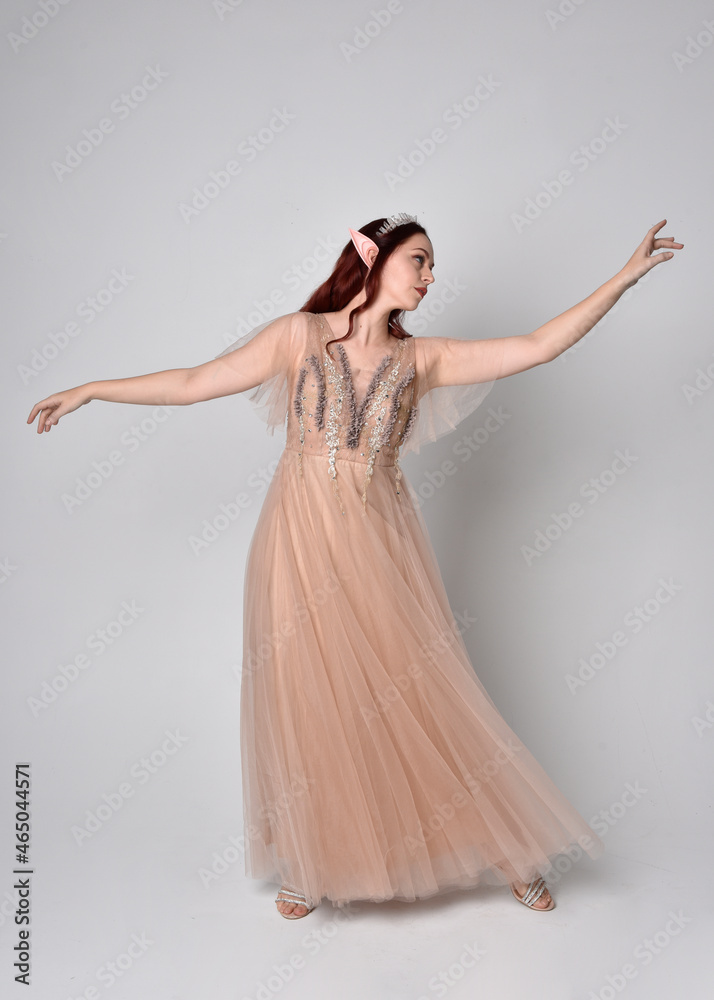Fototapeta premium Full length portrait of red haired girl wearing a creamy fantasy gown and crystal crown, like a fairy goddess costume. standing pose with elegant gestural arm movement , isolated on light studio b