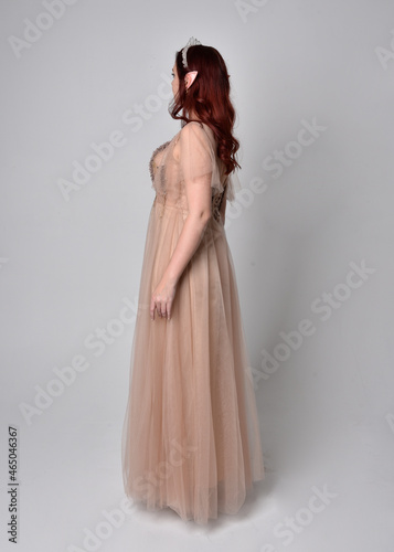 Full length portrait of red haired girl wearing a creamy fantasy gown and crystal crown, like a fairy goddess costume. standing pose with elegant gestural arm movement , isolated on light studio b