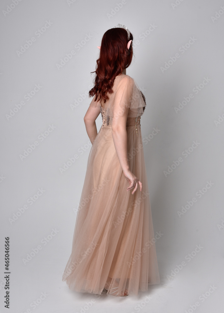 Fototapeta premium Full length portrait of red haired girl wearing a creamy fantasy gown and crystal crown, like a fairy goddess costume. standing pose with elegant gestural arm movement , isolated on light studio b