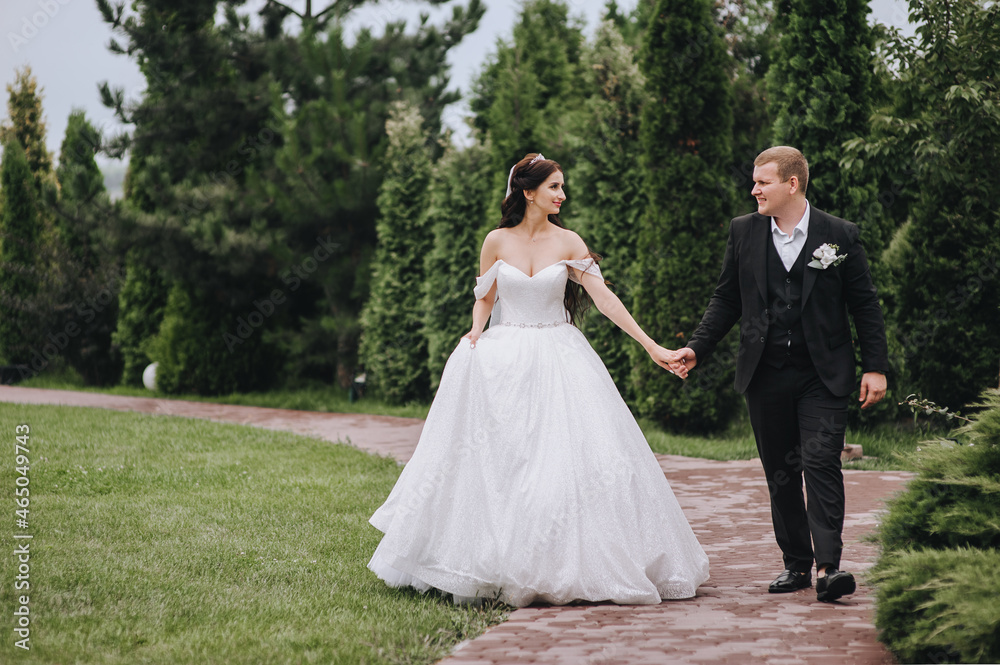 A young groom in a black suit and a beautiful brunette bride in a white dress are walking through the garden, park, road in nature with green plants, holding hands. Wedding portrait of the newlyweds.