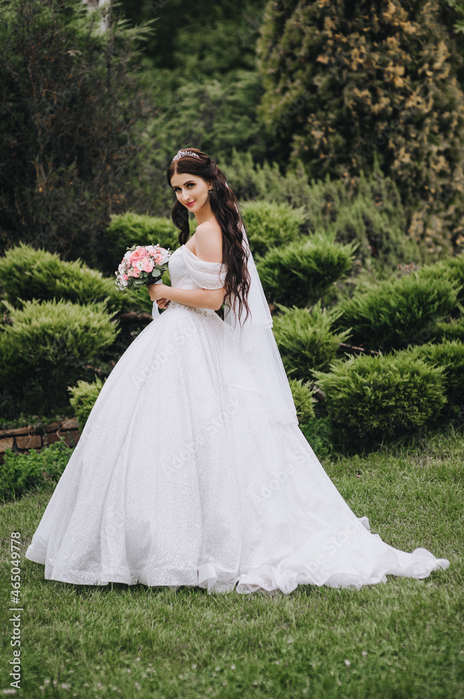 A beautiful bride in a long white dress stands in the garden in nature, against the background of green plants. Wedding photography, portrait.