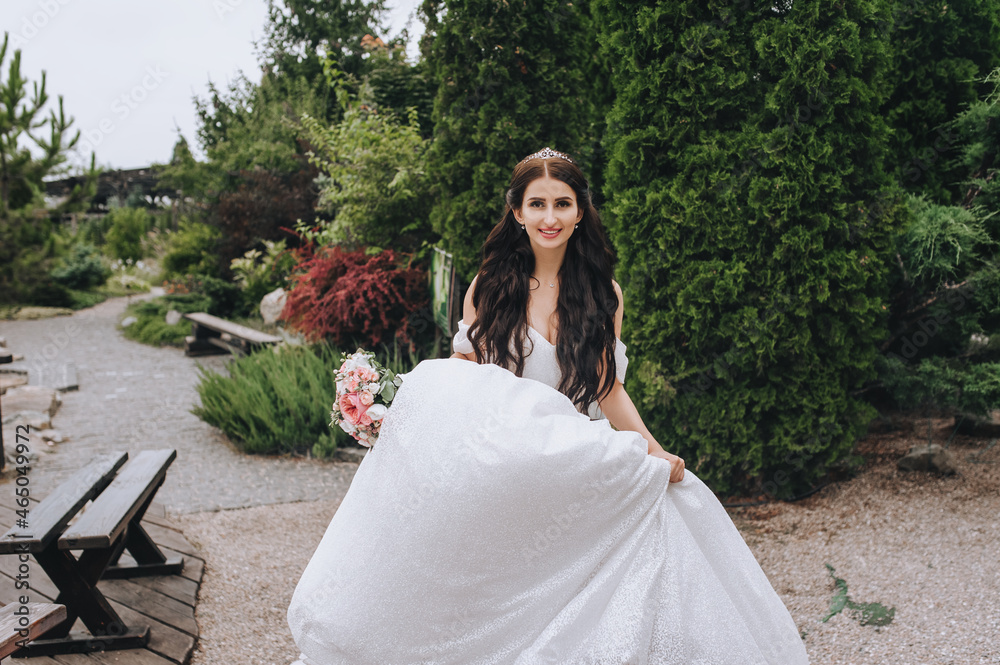 A beautiful, smiling bride in a white dress walks, runs through the garden with green plants. Wedding photography.