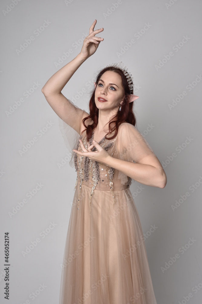 Full length portrait of red haired girl wearing a creamy fantasy gown and crystal crown, like a fairy goddess costume. standing pose with elegant gestural hands, isolated on studio background Full length portrait of red haired girl wearing a creamy fantasy gown and crystal crown, like a fairy goddess costume. standing pose with elegant gestural hands, isolated on studio background