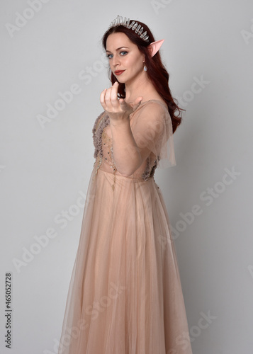 Full length portrait of red haired girl wearing a creamy fantasy gown and crystal crown, like a fairy goddess costume. standing pose with elegant gestural hands, isolated on studio background