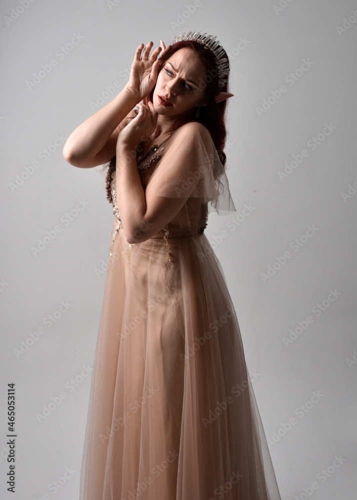 Full length portrait of red haired girl wearing a creamy fantasy gown and crystal crown, like a fairy goddess costume. standing pose with elegant gestural hands, isolated on studio background Full length portrait of red haired girl wearing a creamy fantasy gown and crystal crown, like a fairy goddess costume. standing pose with elegant gestural hands, isolated on studio background