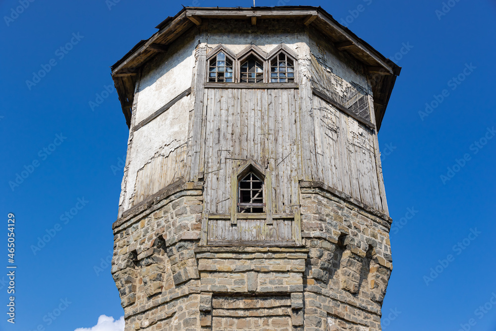 Obraz premium The upper part of an ancient watchtower made of stone blocks and wood paneling with windows against the blue sky on a bright sunny summer day. Close-up