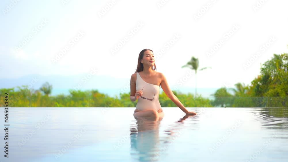 Charming and sexy young Asian woman strokes hair relaxing in infinity pool with legs crossed and holding sunglasses. Slow-motion Charming and sexy young Asian woman strokes hair relaxing in infinity pool with legs crossed and holding sunglasses. Slow-motion