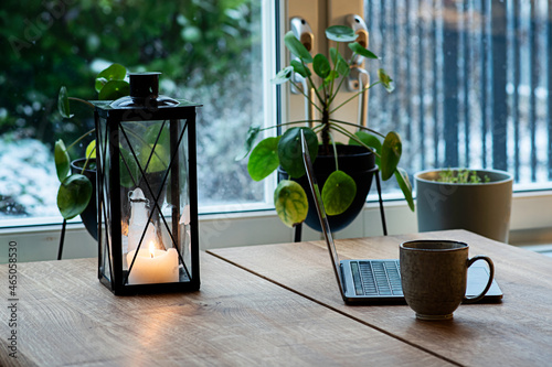 Remote work with laptop and coffee during winter season with candle lantern on wooden table to create cozy atmosphere indoors.