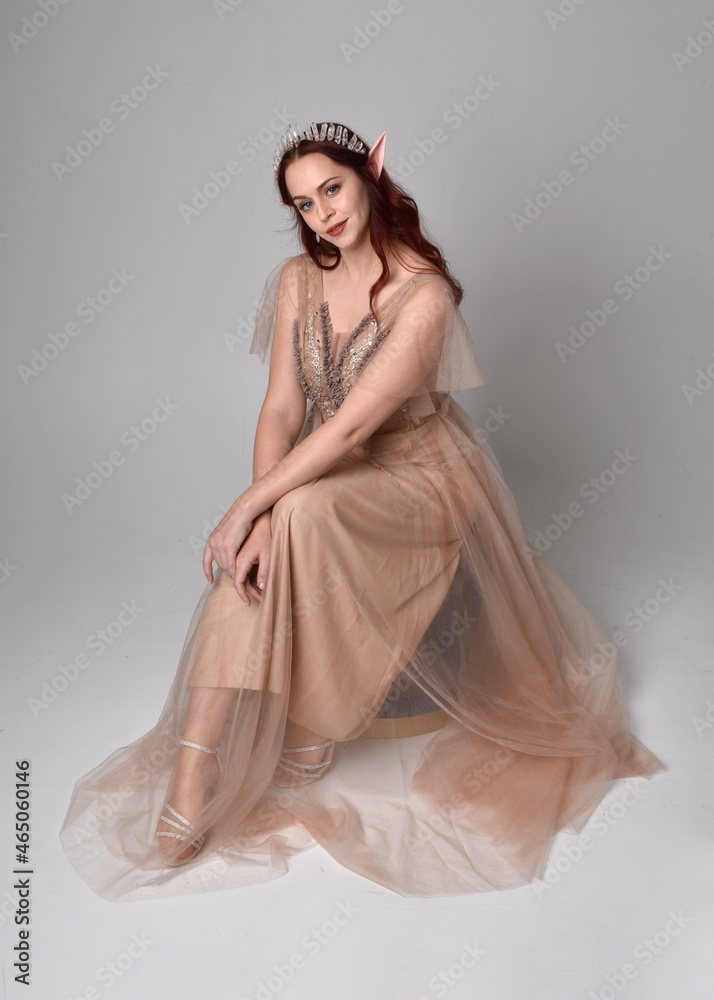Full length  portrait of red haired  girl wearing a creamy fantasy gown and crystal crown, like a fairy goddess costume.  sitting pose with elegant gestural hands, isolated on light studio background.