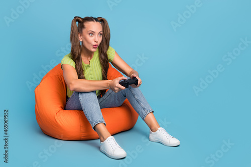 Full length photo of funky young brunette lady sit on chair play station wear jeans t-shirt sneakers isolated on blue color background