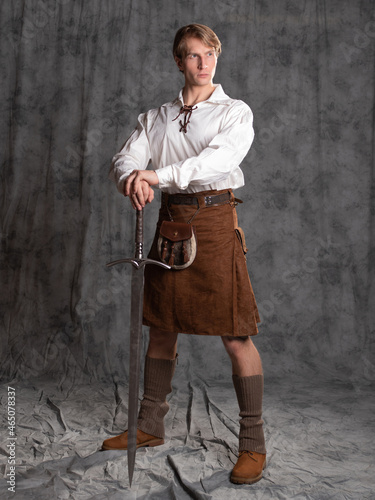 A young man in a leather kilt and a white lace-up blouse. A Scottish knight with a two-handed sword. Full-length Photo in the studio on a gray background