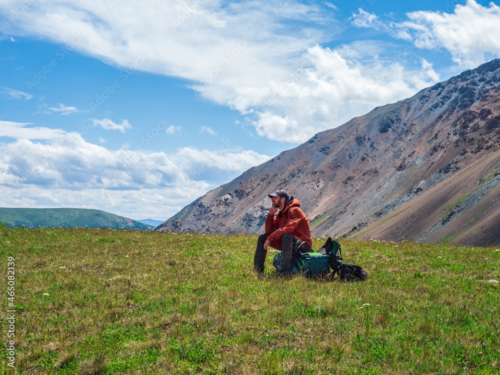 Fototapeta premium Tired tourist sits on a large backpack and happily looks into the distance against the background of blue dramatic clouds against the background of tundra mountains. Rest time on a hike, a rest stop.