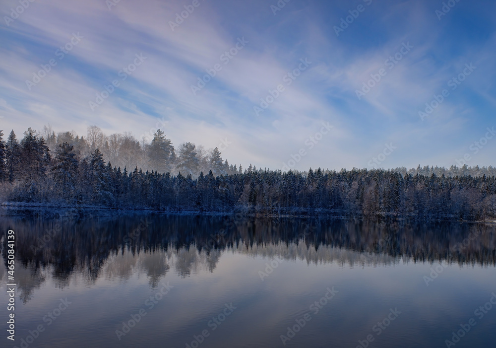 a snow-covered forest with the first snow, a reflection in the lake against the background of a blue sky with clouds