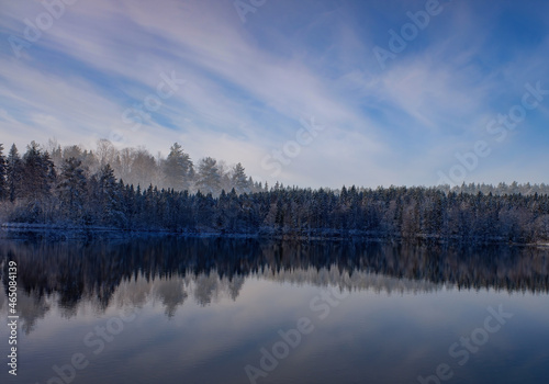 a snow-covered forest with the first snow, a reflection in the lake against the background of a blue sky with clouds © Dmitry